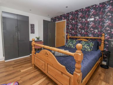 A bedroom with a wooden bed with patterned bedding and pillows a floral wallpaper wall two grey wardrobes wooden flooring and a small desk with a chair at The Old Station House in Withernsea