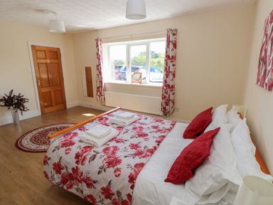A bedroom with a bed with red and white floral bedding two red pillows a window with floral curtains a wooden door and a round rug on a wooden floor at Hillside Lodge in Newtown