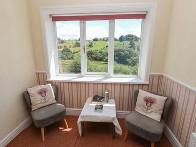 A small sitting area with two gray chairs with floral cushions and a table with books by a window showing green hills at Hillside Lodge in Newtown