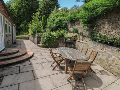 An outdoor patio with a wooden table and chairs a barbecue and stone walls surrounded by greenery at Hillside Lodge in Newtown