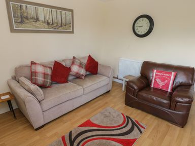 A living room with a beige sofa and a brown leather armchair with red cushions a wall clock and framed forest picture at Hillside Lodge in Newtown