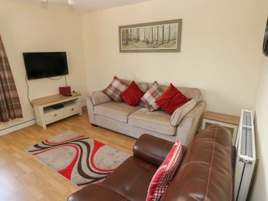 A living room with a beige sofa and a brown leather armchair a wall-mounted TV a wooden TV stand a patterned rug and a painting of a forest at Hillside Lodge in Newtown