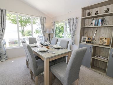 A dining area with six grey chairs around a wooden table by large windows and a shelving unit at Olivias Haven in Wemyss Bay
