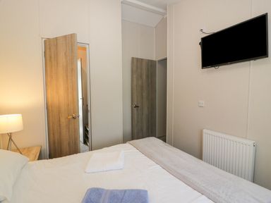 A bedroom with a bed a towel on the bed a bedside table with a lamp two open wooden doors a wall mounted television and a radiator at Olivia's Haven in Wemyss Bay