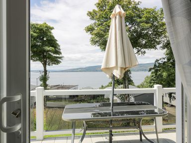 A balcony with a glass table and closed umbrella overlooking a body of water and trees at Olivia's Haven in Wemyss Bay