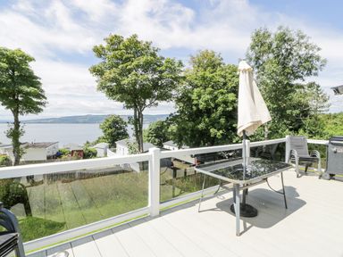 An outdoor deck with a glass table umbrella chairs and a grill overlooking trees and water at Olivias Haven in Wemyss Bay