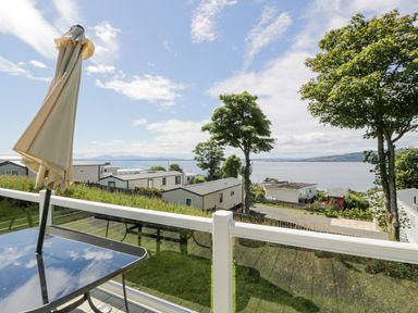 A balcony with a glass table and closed umbrella overlooking caravans trees and a body of water at Olivias Haven in Wemyss Bay