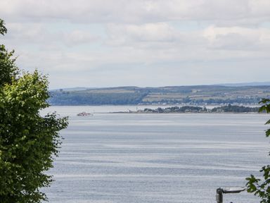 A coastal view with water, trees in the foreground, and land with buildings and hills in the background at Olivia's Haven in Wemyss Bay
