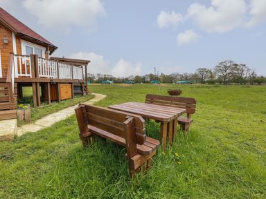 An outdoor wooden table with benches on grass near a wooden house with a porch at Dragonfly Retreat in Upper Marham
