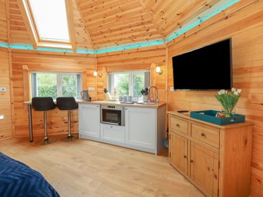 A wooden kitchen interior with bar stools sink microwave TV and a cabinet with flowers on a tray at Dragonfly Retreat in Upper Marham