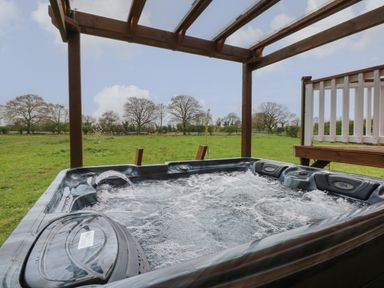 A bubbling outdoor hot tub under a wooden pergola with a grassy field and trees in the background at Dragonfly Retreat in Upper Marham
