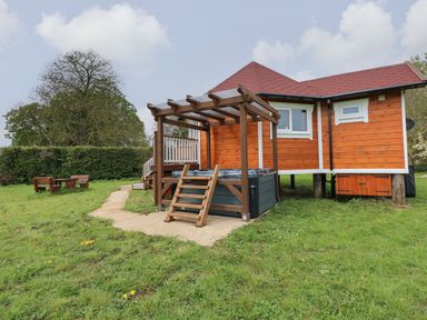 A wooden cabin with a hot tub under a pergola and outdoor seating on a grassy lawn at Dragonfly Retreat in Upper Marham
