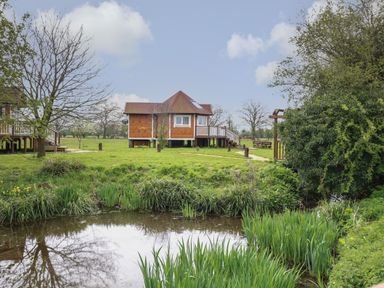 A wooden cabin on stilts near a small pond with grass and trees at Dragonfly Retreat in Upper Marham