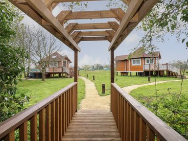 A wooden pergola and bridge over a path leading to two elevated wooden cabins in a grassy area at Dragonfly Retreat in Upper Marham