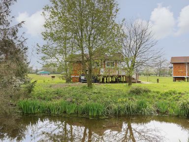 A pond with grass and trees in front of wooden cabins in a field at Dragonfly Retreat in Upper Marham