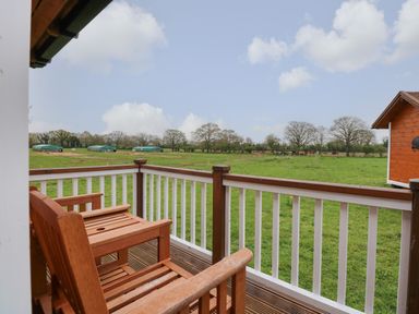 A wooden balcony with two chairs overlooking a grassy field with trees and small structures at Honeybee Retreat in Pentney near Upper Marham