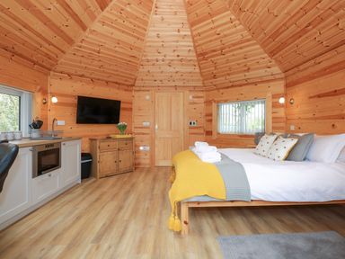 A bedroom with wooden walls and ceiling a bed with pillows and folded towels a kitchenette with a sink microwave and cabinets a wooden cabinet with a vase of flowers a wall-mounted television at Honeybee Retreat in Pentney near Upper Marham