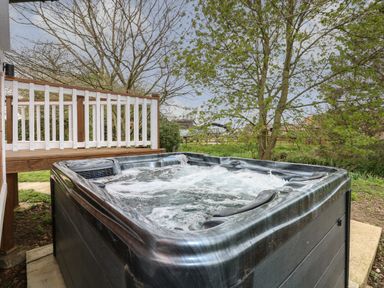 An outdoor hot tub with bubbling water next to a wooden deck and trees at Honeybee Retreat in Pentney near Upper Marham