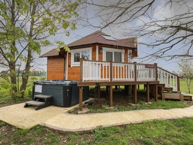 A wooden cabin with a white railing deck and a hot tub on a concrete path surrounded by grass and trees at Honeybee Retreat in Pentney near Upper Marham