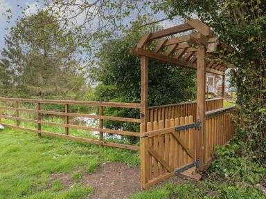 A wooden gate with an arched pergola and fence next to a river in a garden at Honeybee Retreat in Pentney near Upper Marham