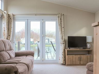 A living room with a sofa a television on a wooden cabinet and glass doors with curtains opening to an outside deck with chairs at Tiptoes Lodge in Carnforth