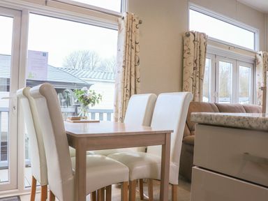 A dining area with a wooden table and four white chairs next to large windows with patterned curtains at Tiptoes Lodge Carnforth