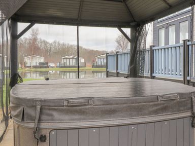 A covered outdoor hot tub next to a fenced deck overlooking a pond with houses and trees in the background at Tiptoes Lodge in Carnforth