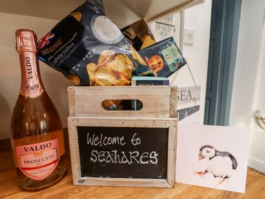 A wooden crate with snacks a bottle of Valdo prosecco and a card with a puffin illustration at Seahares in Seahouses