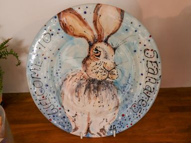A decorative plate with a painted rabbit and the text welcome to seahares on a wooden surface