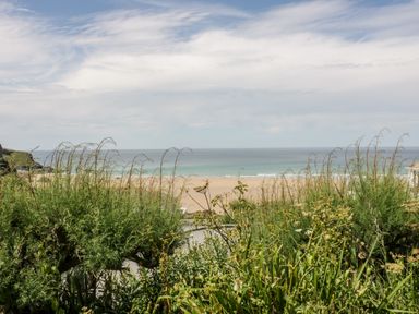 A coastal view with green vegetation in the foreground and a sandy beach meeting the ocean under a partly cloudy sky at Clematis House in St Columb Major