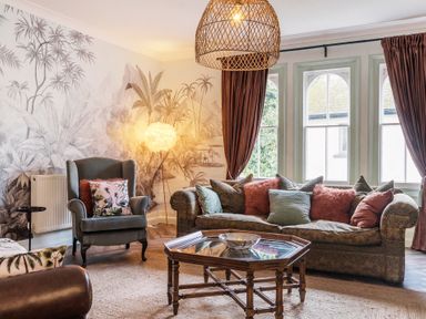 A living room with patterned sofa and armchair with cushions a wooden coffee table and large windows with curtains at Clematis House in St Columb Major
