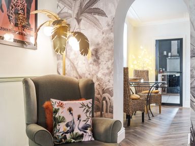 A sitting area with an armchair with pillows and a nearby lamp shaped like palm leaves next to an archway leading to a dining area with woven chairs and a glass table at Clematis House in St Columb Major