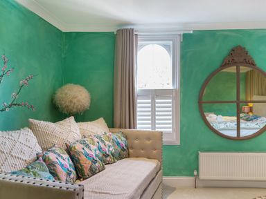 A sitting area with a beige couch with decorative pillows a round mirror a window with shutters and beige curtains and a radiator at Clematis House in St Columb Major