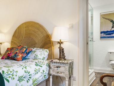 A bedroom with a floral bedspread a wicker headboard a bedside table with a lamp and a glimpse of an adjoining bathroom at Clematis House in St Columb Major