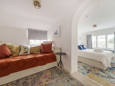 A small seating area with cushions and a side table next to a bedroom with a bed and windows at Clematis House in St Columb Major