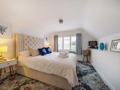 A bedroom with a bed with pillows and folded towels a nightstand with a lamp a window with curtains and a television on a shelf at Clematis House in St Columb Major