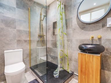 A bathroom with a toilet a shower with glass partition and a wooden vanity with a black sink and a round mirror at Clematis House in St Columb Major