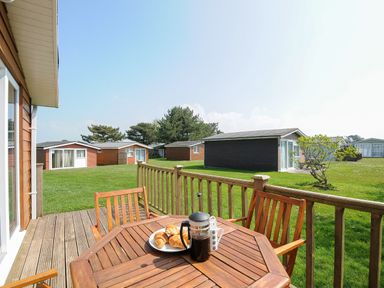 A wooden deck with table and chairs overlooking green grass and small cottages at Little Trebah Atlantic Bays Holiday Park near St Merryn