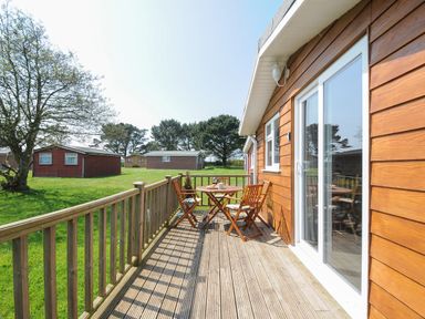 A wooden deck with a table and chairs outside wooden cabins on a grassy area at Little Trebah in Atlantic Bays Holiday Park near St Merryn
