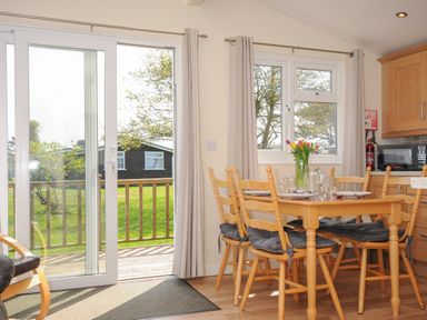 A dining area with a wooden table and chairs set with plates and glasses next to sliding glass doors and windows at Little Trebah Atlantic Bays Holiday Park near St Merryn