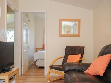 A living room with black leather chairs with orange cushions a wooden TV stand with a TV and a mirror on the wall at Little Trebah Atlantic Bays Holiday Park near St Merryn
