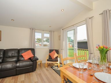 A living room with a black leather sofa a chair with orange cushions windows and a dining table with place settings and flowers at Little Trebah Atlantic Bays Holiday Park near St Merryn