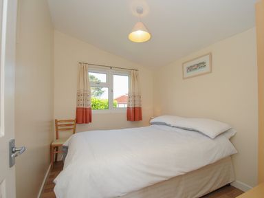 A bedroom with a double bed a wooden chair a window with curtains and a framed picture at Little Trebah Atlantic Bays Holiday Park near St Merryn
