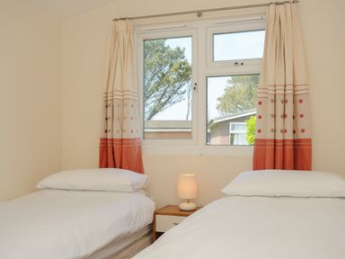 A bedroom with two single beds separated by a nightstand with a lamp and a window with curtains at Little Trebah Atlantic Bays Holiday Park near St Merryn