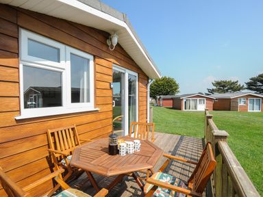 A wooden outdoor patio with table and chairs next to a wooden cabin at Little Trebah Atlantic Bays Holiday Park near St Merryn