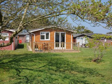 A wooden cabin with a small deck and outdoor table set on grass with trees at Little Trebah Atlantic Bays Holiday Park near St Merryn
