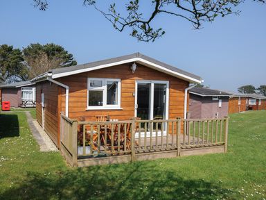 A wooden cabin with a small deck and outdoor furniture on a grassy area at Little Trebah Atlantic Bays Holiday Park near St Merryn