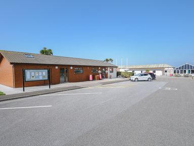 A parking area in front of wooden and white buildings with cars and a notice board at Little Trebah Atlantic Bays Holiday Park near St Merryn