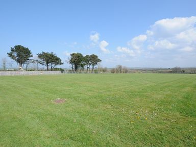 A large grassy field with trees and a fence under a partly cloudy sky at Little Trebah Atlantic Bays Holiday Park near St Merryn