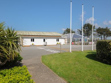 An outdoor area with a white building a glass pavilion flagpoles grass and bushes at Little Trebah Atlantic Bays Holiday Park near St Merryn